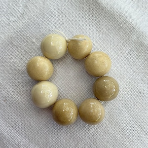 Peut inclure: Un arrangement circulaire de huit perles sphériques enfilées sur un fin cordon blanc. Les perles présentent une finition brillante avec des nuances variées de crème, beige et brun clair, affichant une texture de surface lisse et polie.