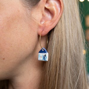 Può includere: Orecchini a cerchio dorati con un piccolo ciondolo a forma di casa in ceramica bianca. La casa ha un tetto blu ed è decorata con motivi floreali blu e rossi. Indossati da una persona con i capelli biondi.