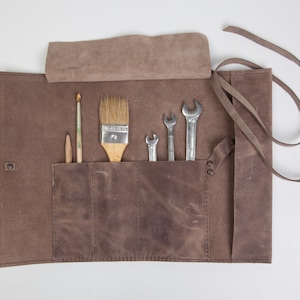 May include: A brown leather roll-up tool holder with pockets for tools and brushes. The holder has a strap for carrying and is open to reveal the tools inside. The tools include wrenches and paint brushes.