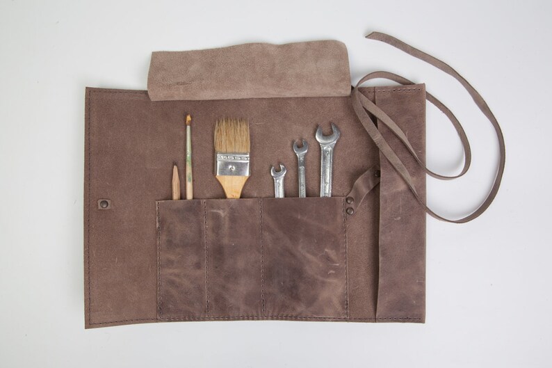 May include: A brown leather roll-up tool holder with pockets for tools and brushes. The holder has a strap for carrying and is open to reveal the tools inside. The tools include wrenches and paint brushes.