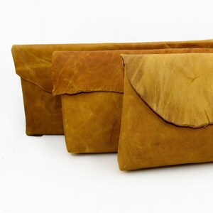 May include: Three brown leather clutches with a flap closure. The clutches are arranged in a row, with the largest clutch in the back and the smallest clutch in the front.