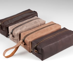 May include: Four rectangular leather pencil cases in shades of brown, tan, and gray are arranged on a white surface. Each case has a zipper closure and a matching leather pull tab. The cases vary in color and texture.