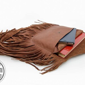May include: A brown leather fringe bag with a flap closure and a pocket on the front. The bag is open and has a red notebook, a black phone, and a brown leather notebook inside.