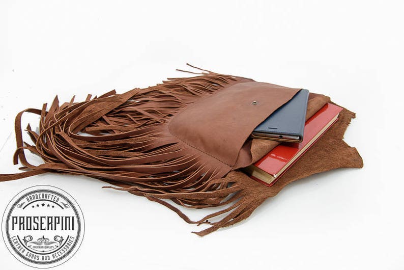 May include: A brown leather fringe bag with a flap closure and a pocket on the front. The bag is open and has a red notebook, a black phone, and a brown leather notebook inside.