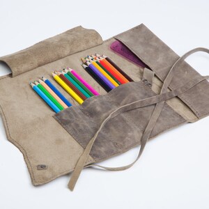 May include: A brown leather pencil roll with a strap closure. The roll is open and filled with colored pencils.