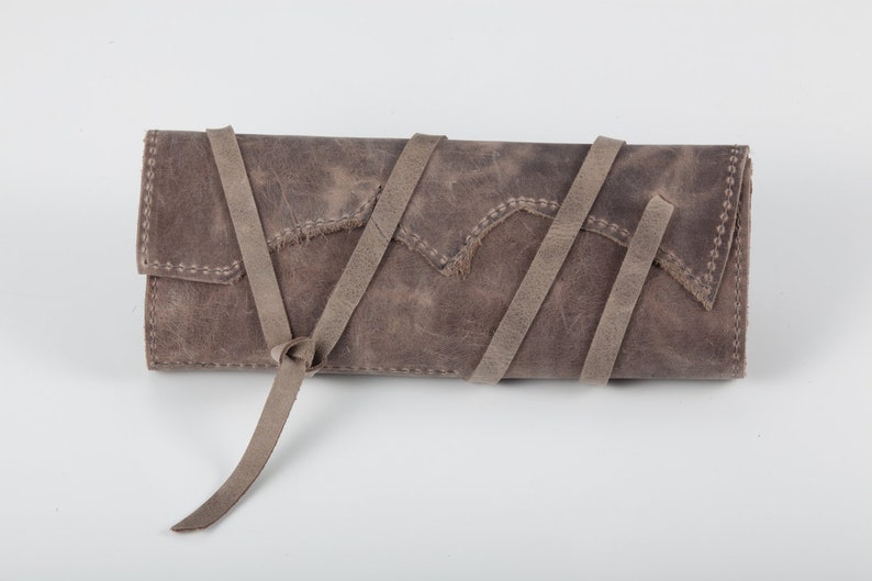 May include: A gray leather clutch with a long strap that can be tied in a knot. The clutch has a distressed look and is made of a soft, supple leather.