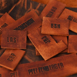 May include: A collection of brown leather labels with various text, including "M. GUILLOU", "BROOMSWAN", "LAS", "CRP", "BSH", "PVC", "PFEFFENBERGER", "MERLOS", "DGS", and "PLUKYS".