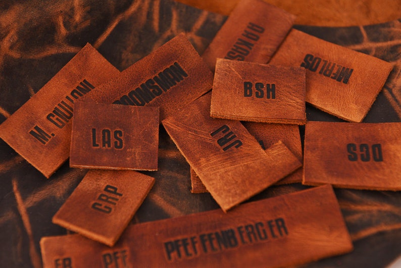 May include: A collection of brown leather labels with various text, including "M. GUILLOU", "BROOMSWAN", "LAS", "CRP", "BSH", "PVC", "PFEFFENBERGER", "MERLOS", "DGS", and "PLUKYS".