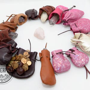 Peut inclure: Une collection de petites pochettes en cuir de différentes couleurs et styles, notamment rose, marron, or et noir. Certaines pochettes sont décorées de paillettes ou de pièces de monnaie. Les pochettes sont disposées en cercle autour d'un cristal rose.