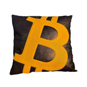 Puede incluir: Cojín cuadrado negro con un gran símbolo Bitcoin dorado. El símbolo Bitcoin está centrado en el cojín. El cojín tiene un aspecto texturizado.