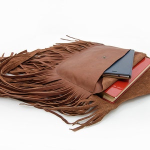 May include: A brown leather fringe bag with a flap pocket and a zipper closure. The bag is open and has a red notebook and a black tablet inside.