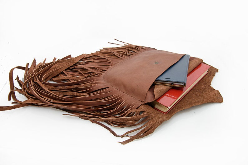 May include: A brown leather fringe bag with a flap pocket and a zipper closure. The bag is open and has a red notebook and a black tablet inside.