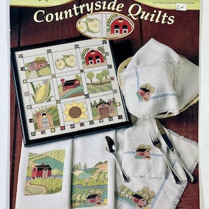 May include: A book titled "Countryside Quilts" with 6 fun designs for table linens and home decor. The cover features a colorful illustration of a quilt with a farmhouse, sunflowers, and corn.