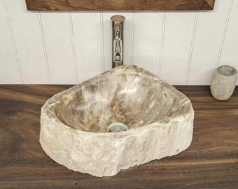 Könnte beinhalten: Ein Aufsatzwaschbecken aus Naturstein mit rauer, strukturierter Außenseite und poliertem Innenbecken, montiert auf einem dunkelbraunen Waschtisch aus Massivholz mit zwei offenen Ablagen, einem Chrom-Wasserhahn und einem weißen Handtuch.