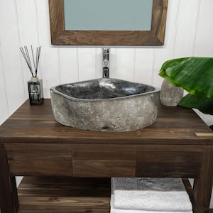 Peut inclure: Un lavabo en pierre grise avec une finition naturelle et texturée est posé sur une vanité en bois brun foncé. L'évier a une forme ovale. Un miroir avec un cadre en bois est suspendu au-dessus de l'évier. Une pile de serviettes blanches et grises est sur l'étagère en dessous.