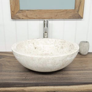 Peut inclure: Une vasque ronde en pierre beige clair mouchetée avec une finition polie. Le bassin présente un intérieur et un extérieur lisses et incurvés, mettant en valeur les variations naturelles de la pierre dans des tons crème et beige.