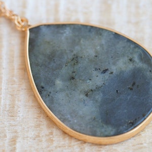 Puede incluir: Un collar con un colgante en forma de lágrima, con una piedra gris oscura engastada en un marco de metal dorado.