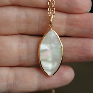 May include: A gold chain necklace with a white iridescent oval pendant. The pendant is set in a gold frame and has a subtle shimmer.