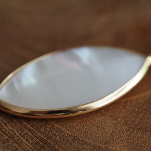 May include: A white mother of pearl pendant with a gold rim. The pendant is shaped like a teardrop and is hanging from a gold chain.