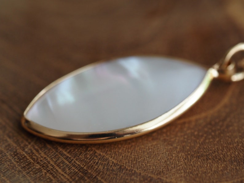 May include: A white mother of pearl pendant with a gold rim. The pendant is shaped like a teardrop and is hanging from a gold chain.