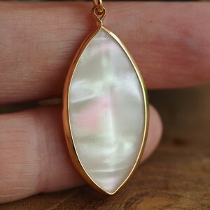 May include: A gold pendant with a white iridescent oval-shaped stone. The stone has a pearlescent finish and a subtle pink hue.