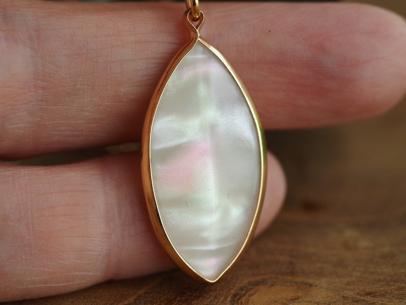 May include: A gold pendant with a white iridescent oval-shaped stone. The stone has a pearlescent finish and a subtle pink hue.