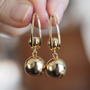 Op de afbeelding: Gouden creolen met een hangende balbedel. De oorbellen zijn gemaakt van glanzend metaal en hebben een eenvoudig ontwerp.