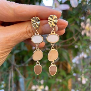 Pode incluir: Brincos pendurados dourados com uma parte superior texturizada e três pedras ovais e em forma de gota descendentes em branco, bege e castanho. Os brincos são segurados contra um fundo floral verde e rosa desfocado.