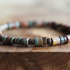 May include: A beaded bracelet with a variety of earth-toned colors, including brown, green, and yellow. The beads are small and cylindrical, strung together to form a circular shape. The bracelet is resting on a wooden surface.