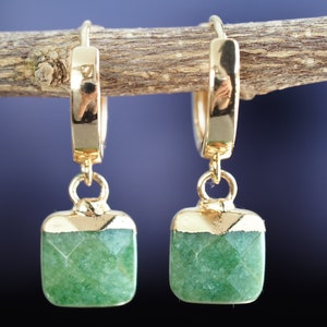 Puede incluir: Pendientes de aro de oro con un colgante cuadrado de piedra verde. Los pendientes están colgados de una rama de madera.