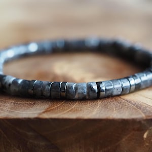 Puede incluir: Un brazalete gris de cuentas con detalles plateados. El brazalete está hecho de pequeñas cuentas redondas y está enhebrado en una cadena plateada.