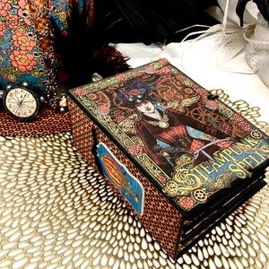 Puede incluir: Un libro de temática steampunk con una cubierta dorada y negra que presenta a una mujer con alas y un reloj. El libro se titula "Steampunk Spells" y está sobre un encaje dorado.