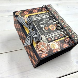 Op de afbeelding: Een decoratieve doos met een ruitpatroon in bruin- en zwarttinten. De doos heeft bloemversieringen, een zwart-wit gestreept lint en de tekst "RISE & SHINE THIS IS MY HAPPY PLACE".