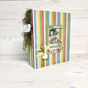 Op de afbeelding: Een decoratief scrapbookalbum met een kleurrijk streepjespatroon in rood, geel, blauw en groen. Het album heeft een ingelijste afbeelding van teddyberen en een konijn, met het woord "celebrate". Een strik en een label met tekst zijn bevestigd.