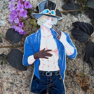 K&ouml;nnte beinhalten: Ein Anime-Pin mit einem Charakter mit blonden Haaren, der einen schwarzen Zylinder mit Brille, eine blaue Jacke, schwarze Shorts und braune Handschuhe tr&auml;gt. Die Figur h&auml;lt eine Pfeife. Der Pin befindet sich vor einem Naturstein-Hintergrund mit lila Blumen.