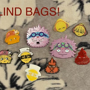 May include: A collection of colorful enamel pins with various character designs. The pins feature a variety of colors, including yellow, pink, orange, and red. The text "BLIND BAGS!" is displayed in red at the top of the image.