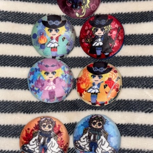 May include: A collection of seven round, colorful buttons featuring anime-style character illustrations. The buttons display various characters in different outfits and color schemes, set against patterned backgrounds. The buttons are arranged on a striped surface.