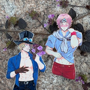 K&ouml;nnte beinhalten: Zwei Aufkleber mit Anime-Charakteren. Ein Aufkleber zeigt einen blonden Charakter in einer blauen Jacke, einer schwarzen Hose und einem Zylinder mit Brille. Der andere Aufkleber zeigt einen pinkhaarigen Charakter in einem blauen Hemd und roten Shorts.