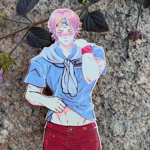 K&ouml;nnte beinhalten: Ein farbenfroher Anime-Pin mit einem m&auml;nnlichen Charakter in einem blauen Hemd und roten Shorts. Der Pin hat ein himmlisches Design auf der Stirn und ein Herzdetail auf den Shorts. Der Charakter hat rosa Haare und posiert auf suggestive Weise.