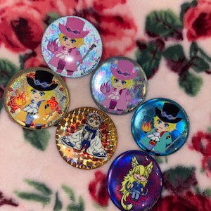 May include: A collection of seven round, colorful pinback buttons featuring anime-style character illustrations. The buttons have holographic and glittery effects, with characters in various outfits and poses. The background is a pink floral pattern.
