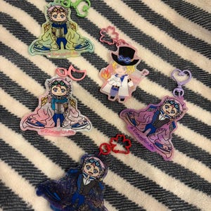 May include: Five acrylic keychains featuring anime-style characters in various outfits. Each keychain has a different colour scheme, including blue, pink, purple, and green. The keychains are attached to various coloured clasps and rings. The background is a striped fabric.