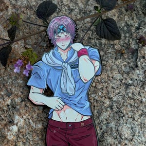 K&ouml;nnte beinhalten: Ein farbenfroher Emaille-Pin mit einem Anime-Charakter mit rosa Haaren, der ein blaues Hemd, bordeauxfarbene Shorts und einen wei&szlig;en Schal tr&auml;gt. Der Pin ist vor einem Naturstein-Hintergrund mit Gr&uuml;n angebracht.