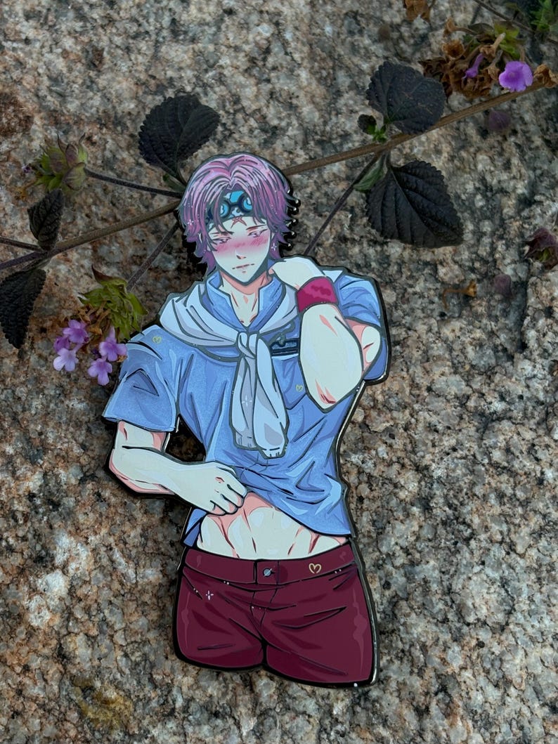 K&ouml;nnte beinhalten: Ein farbenfroher Emaille-Pin mit einem Anime-Charakter mit rosa Haaren, der ein blaues Hemd, bordeauxfarbene Shorts und einen wei&szlig;en Schal tr&auml;gt. Der Pin ist vor einem Naturstein-Hintergrund mit Gr&uuml;n angebracht.