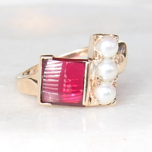 May include: A gold ring featuring a red rectangular stone and three white pearls. The ring is displayed on a white marble surface.