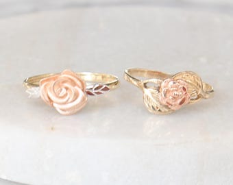 May include: Two gold rings with rose-shaped designs. One ring features a rose with a gold band and silver leaves. The other ring has a rose with gold leaves and a gold band. The rings are on a white surface.