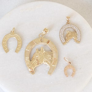 May include: Four gold-tone pendants, including a horseshoe, a horseshoe with a horse head, and two smaller horseshoes. The pendants are on a white marble surface.