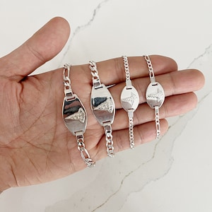 May include: Four silver medical alert bracelets with varying chain designs. Each bracelet has a polished, oval plate bearing a medical symbol. The bracelets are shown on a light-coloured surface.