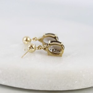 May include: A pair of gold-colored drop earrings with oval-shaped, light brown gemstones. The earrings have a small gold ball at the top and a post for pierced ears. The gemstones are set in a gold bezel setting.
