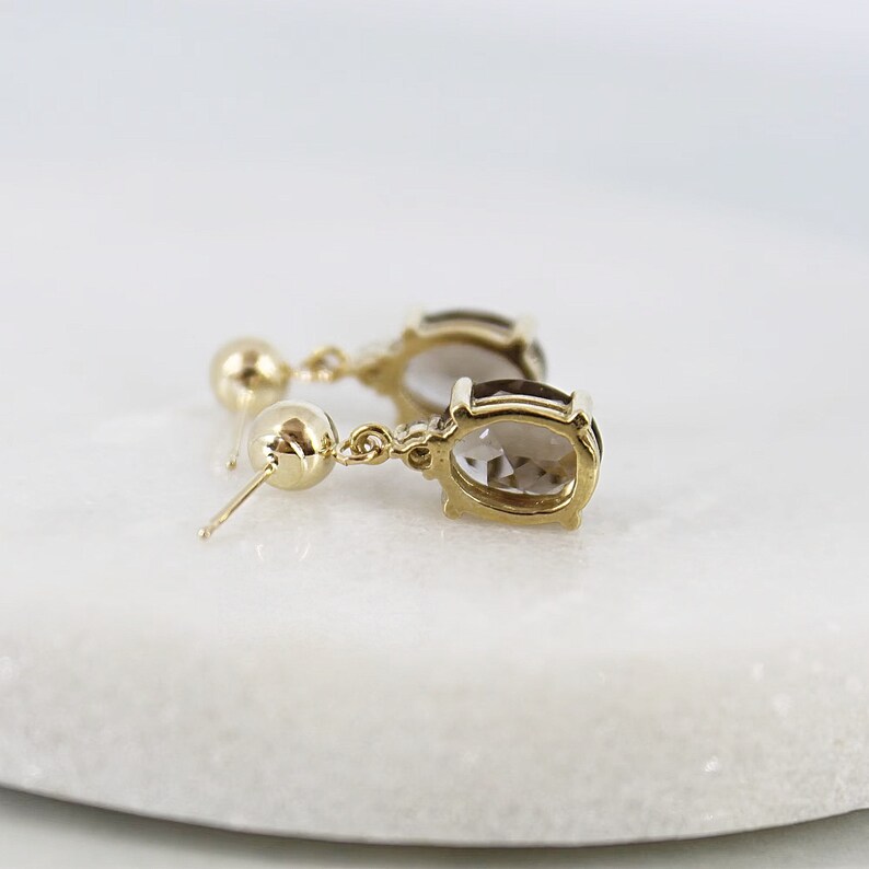 May include: A pair of gold-colored drop earrings with oval-shaped, light brown gemstones. The earrings have a small gold ball at the top and a post for pierced ears. The gemstones are set in a gold bezel setting.