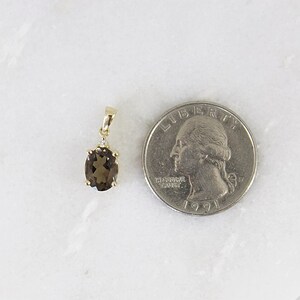 May include: A gold pendant with an oval, smoky quartz gemstone and a small diamond accent. The pendant is next to a U.S. quarter for size comparison. The gemstone is a light brown color.
